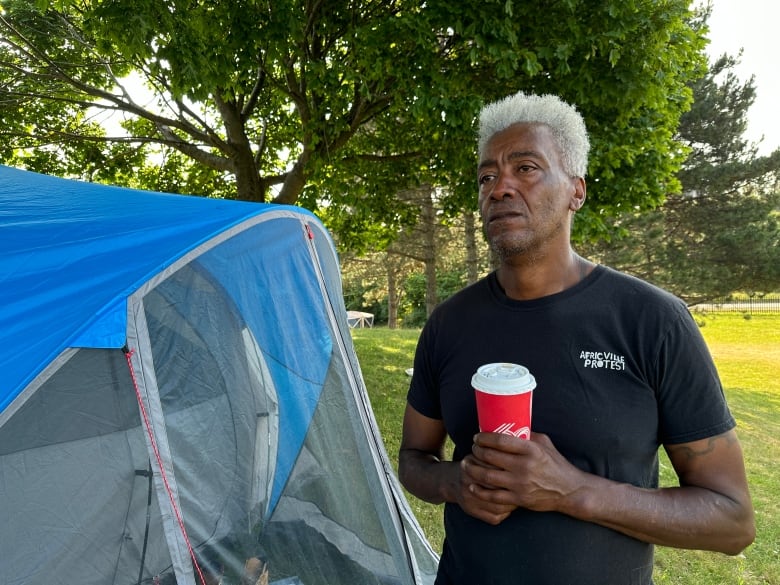 A Black man with white hair, wearing a black t-shirt with a logo that says Africville Protest, stands next to a tent and holds a coffee cup.