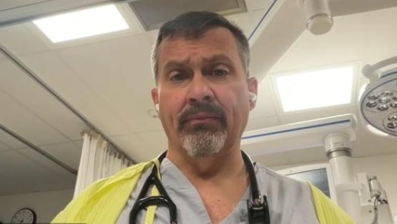 Dr. Paul Parks is wearing grey scrubs and has a stethoscope around his neck as he looks at the camera. Medical equipment and a hospital privacy curtain can be seen behind him