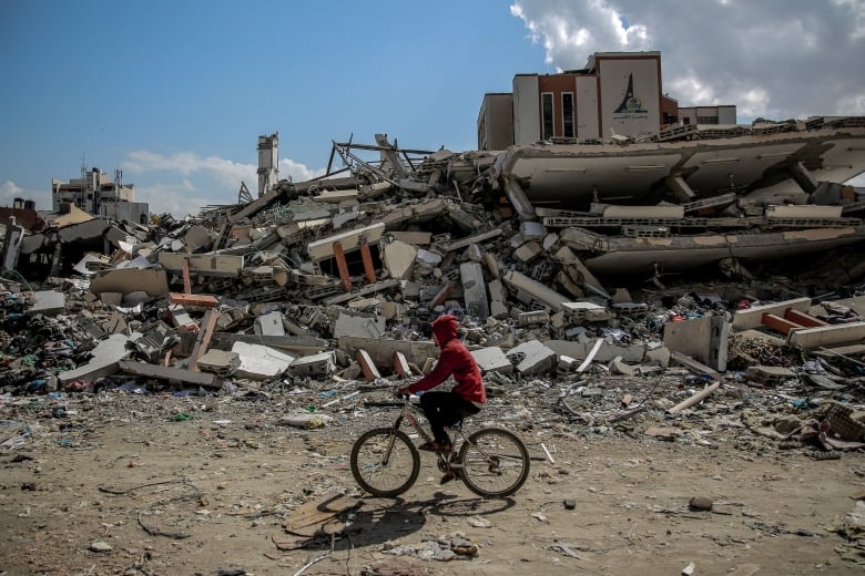 A person rides on a bicycle past the rubble of a building.