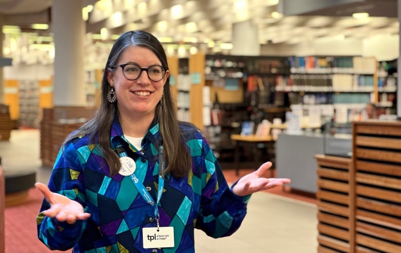 A smiling woman standing in a spacious library space gestures with her hands as she speaks to a person off camera to the right.