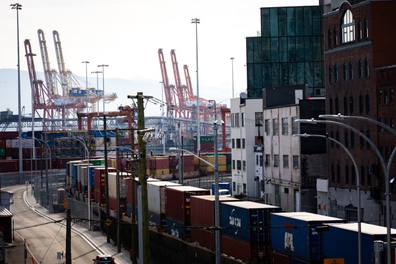 A series of cranes are seen at a port, with cargo containers in the foreground next to buildings.