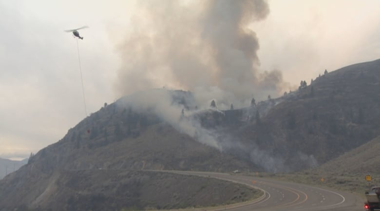 A helicopter is seen flying away from a hilltop that is spewing smoke.