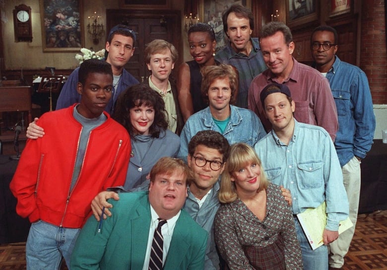 13 SNL actors pose for a photo on the SNL stage. Phil Hartman is located in the top right of the cast