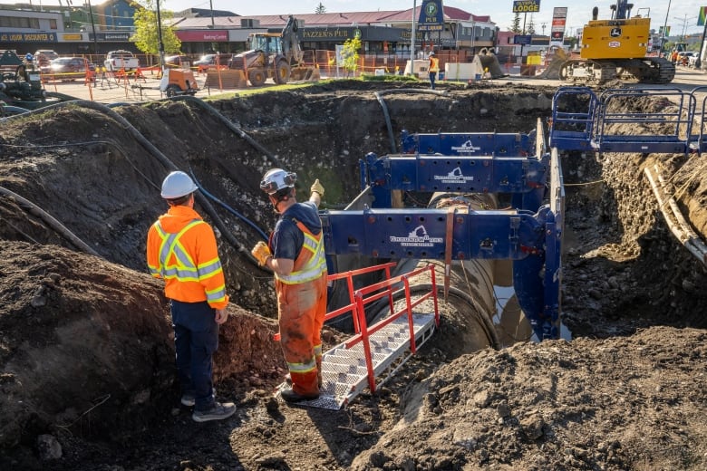 Two workmen stand next to an exposed section of water main pipe.