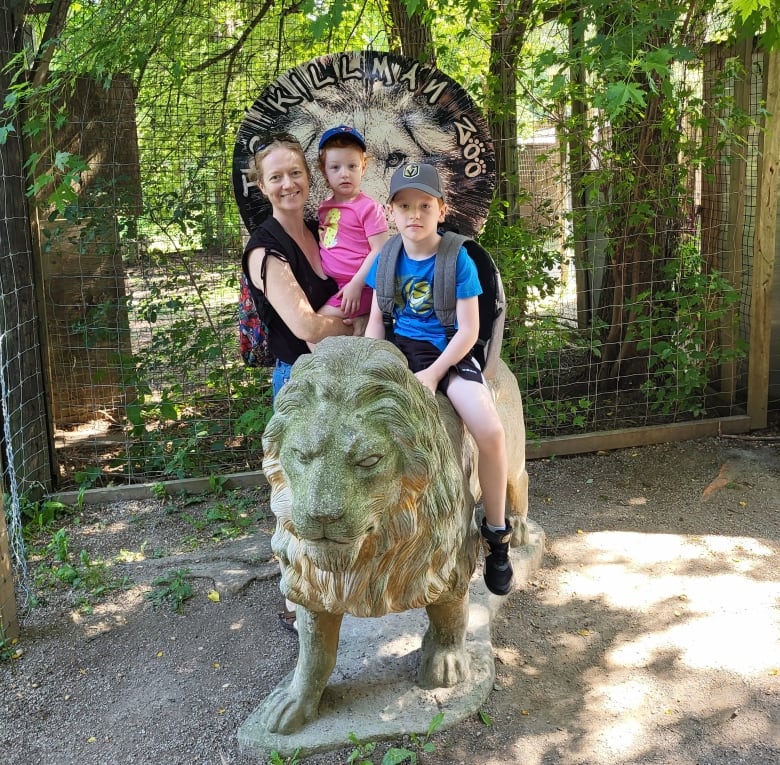 A woman smiles as she holds a young girl next to a boy a few years older who is sitting on a statue of a lion. A sign in the background reads "The Killman Zoo."