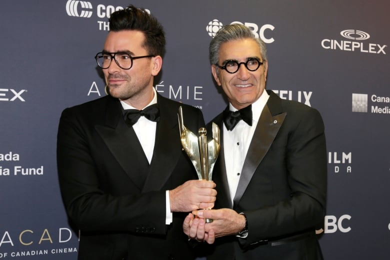 Two men wearing tuxedos hold an award together. 