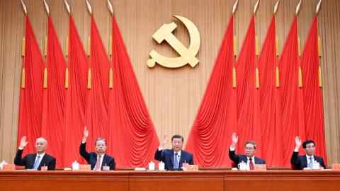 Five Asian men sit behind a podium and raise their right hands, while flanked by ten red flags and a golden sickle and hammer