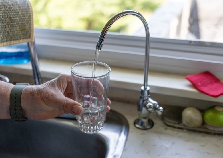 A hand holding a clear glass underneath a small silver tap beside a kitchen sink. Water is coming from the tap into the glass. 