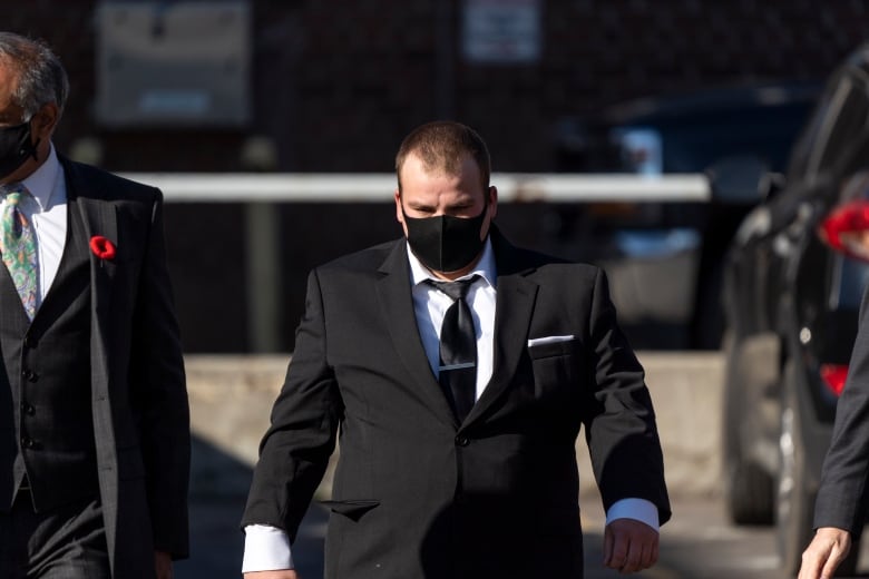 A man in a suit and wearing a mask walks with cameras and media following behind him. 