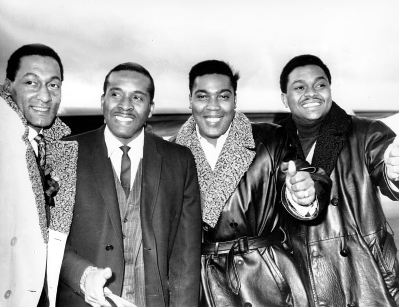 Four dark complected men wearing coats smile while posing for a black and white picture that appears to be decades old.