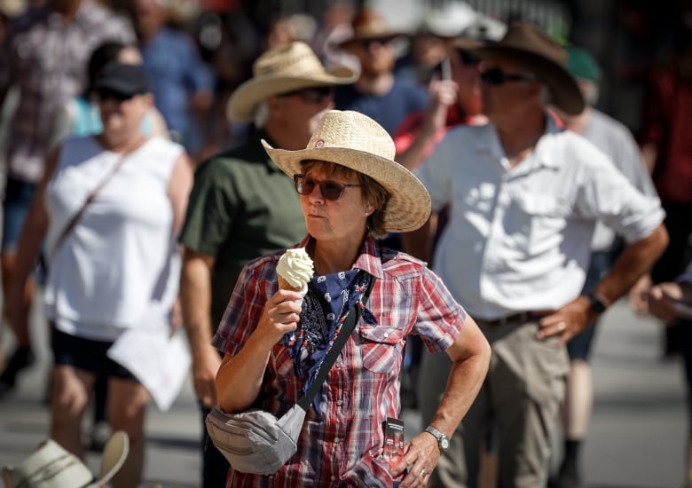 A woman wearing a wide-brimmed hat and sunglasses holds an ice cream cone in a crowd.