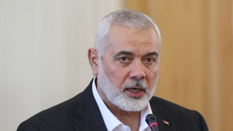 A man with grey hair and a white beard stands at a podium wearing a white shirt and black suit jacket.