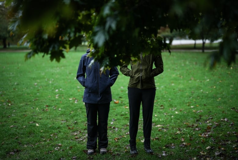 Leaves hanging from a tree cover the faces of two women standing in a park, wearing rain coats.