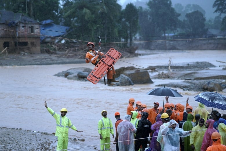 A helmeted man in a mustache is shown travelling above a body of water on a zip-line while carrying a stretcher.