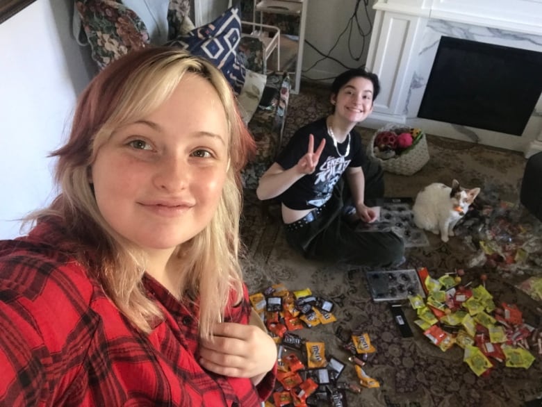 Two smiling teenagers pose for a selfie. One of them is sitting cross-legged on the floor and is surrounded by candy.  