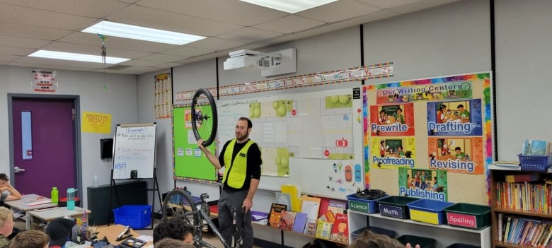 A man in a green safety vest holds up a bike tire in one hand while standing in an elementary school classroom. 