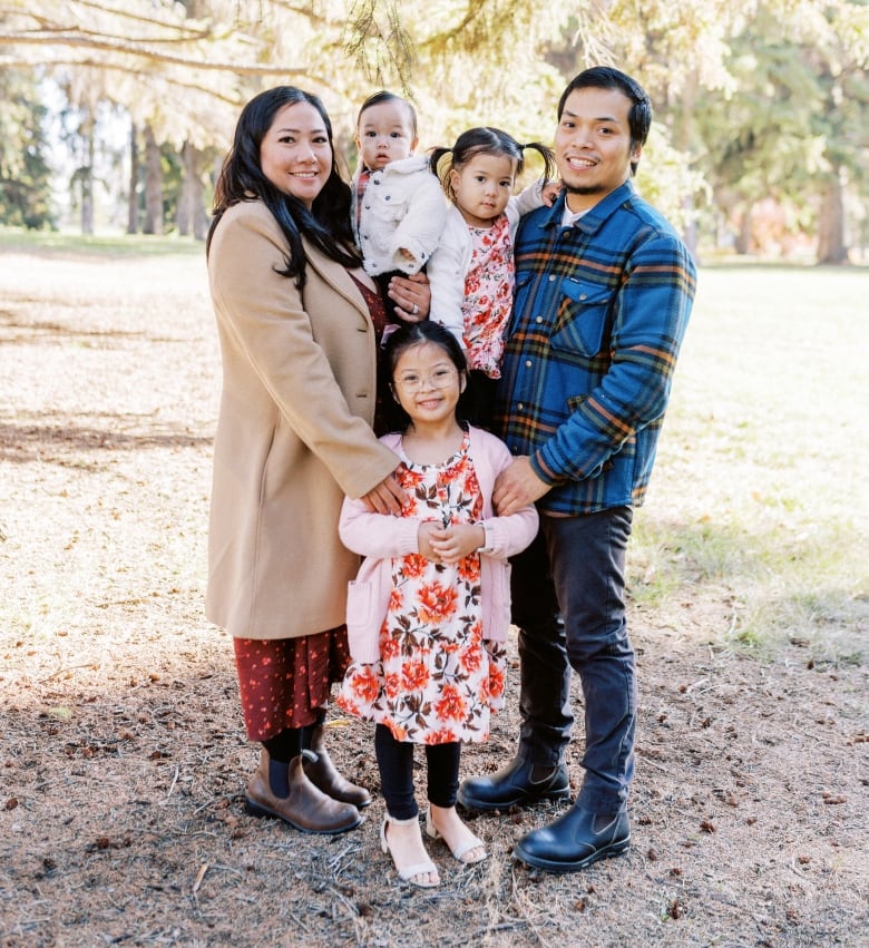 A smiling woman in a tan coat holds a baby, while a man on the right holds another baby. In front of them stands a smiling older girl in a flower dress.