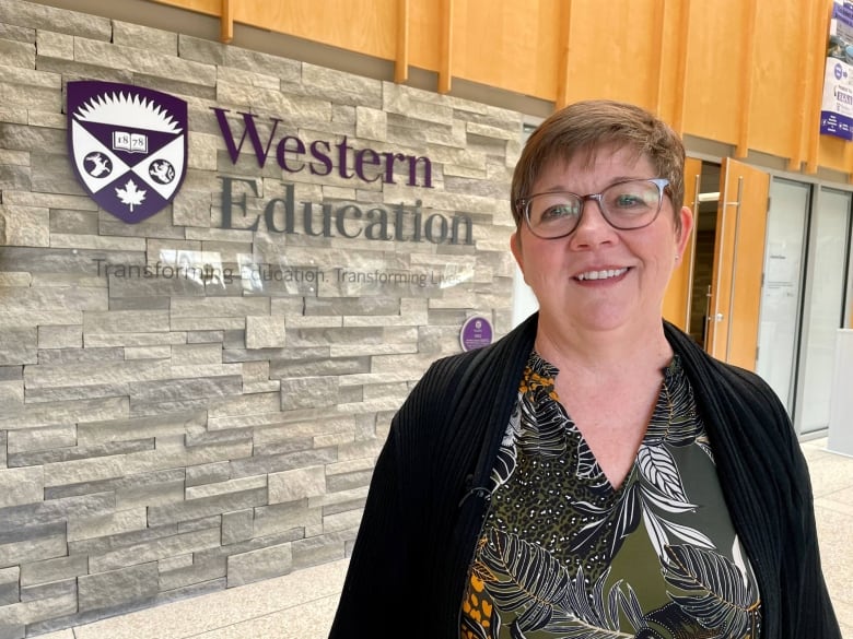 A woman with short brown hair and glasses smiles in front of a brick wall. There is a sign that reads "Western Education." 