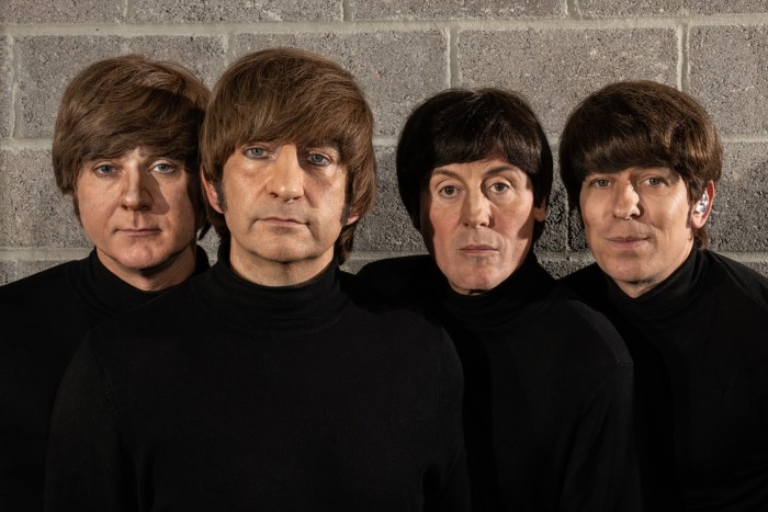 Four men with bowl haircuts wearing black stand against a brick wall