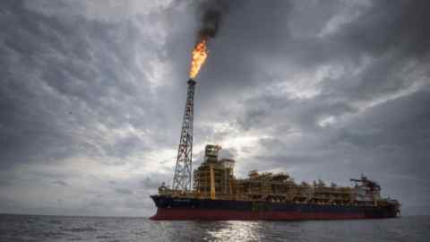 An offshore oil platform in the ocean with a tall flare stack burning off excess gas against a backdrop of a cloudy sky