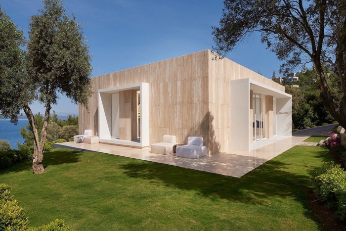 a rectangular minimalist, modern building set in a lush green landscape. The exterior is clad in sand-coloured material