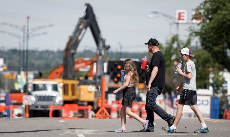 Three people walk in front of a construction scene pictured in the background.