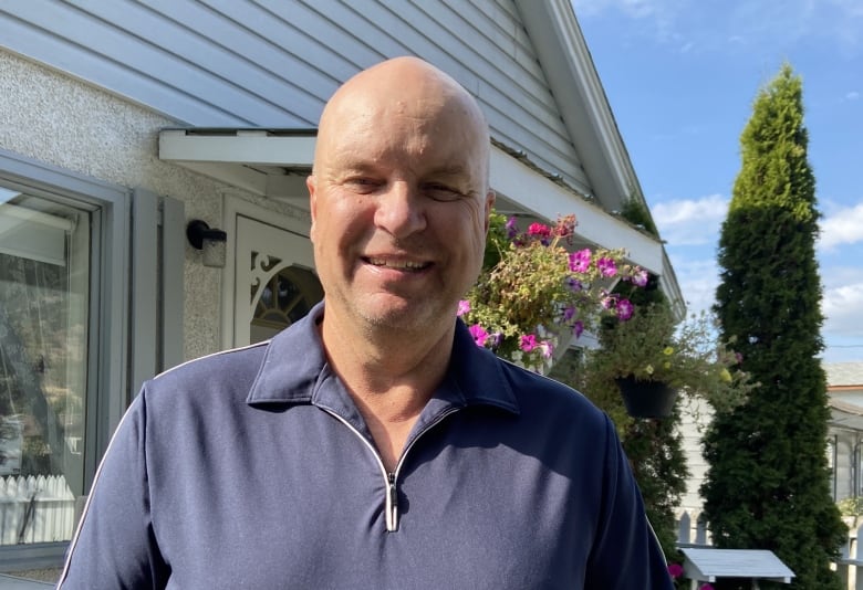 A man in a blue shirt stands in front of a house with flowers in the background. 