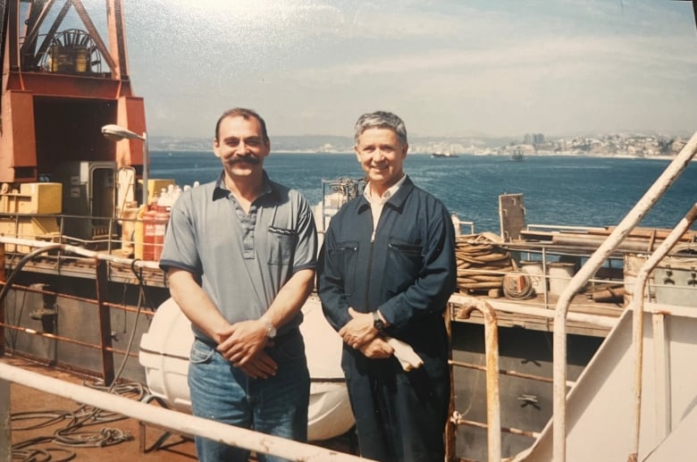 Two smiling men stand on the deck of an oil tanker. 