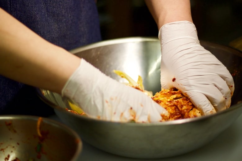 A person wearing white gloves makes kimchi in a metal bowl.