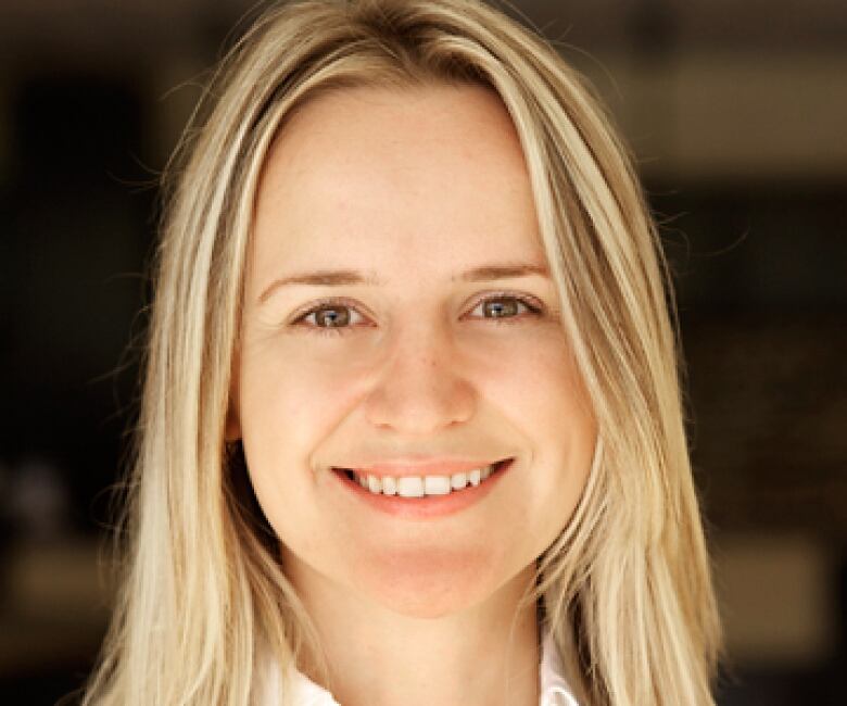 A woman with long blond hair, wearing a white shirt, smiles at the camera.