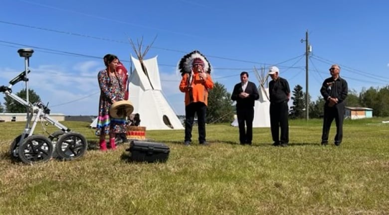 A chief speaks near a piece of equipment in front of two teepees as four other people stand nearby.