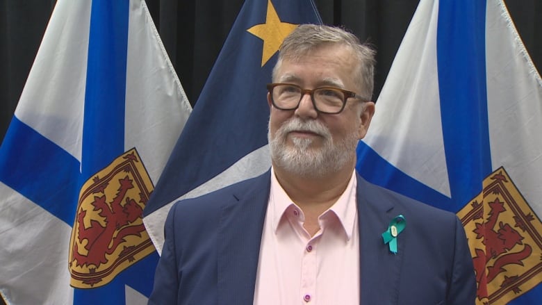 A man wearing glasses and a suit in front of Nova Scotia flags. 