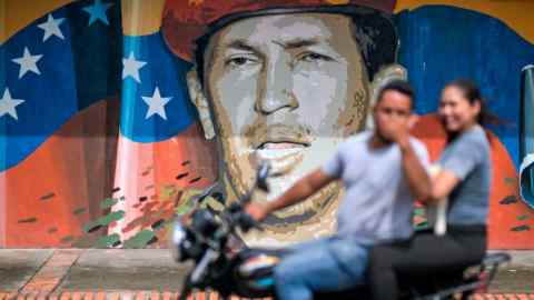 People on a motorbike ride past a mural of mural of Hugo Chávez