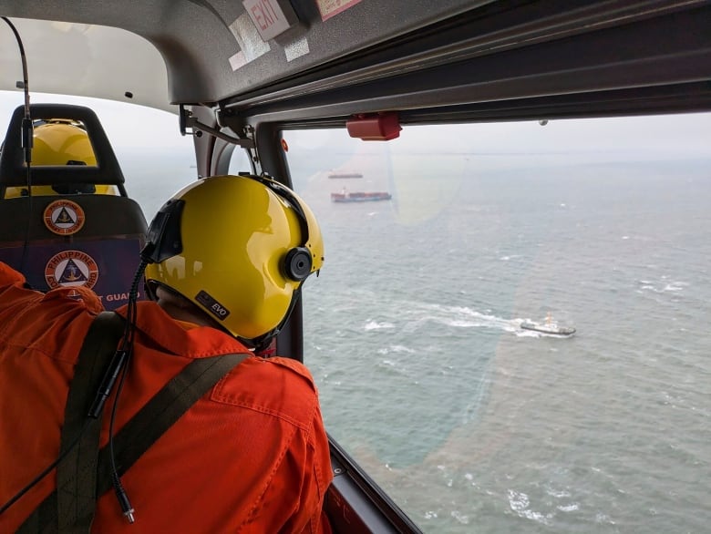 The back of the head of a helmeted person in a jumpsuit is shown, aboard a small plane or helicopter above a large body of water.