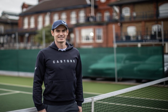 Andy Murray standing and smiling on a tennis court, wearing a hoody and baseball cap