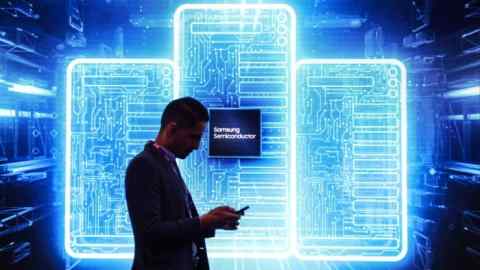 A man uses his phone in front of a large digital display with circuit designs and the words ‘Samsung Semiconductor’