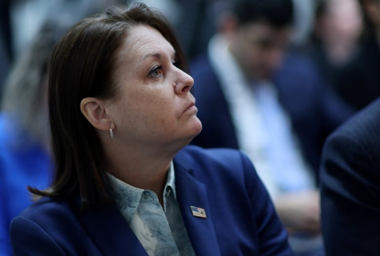 A woman with dark hair past her shoulder wearing a blazer is shown seated in profile.