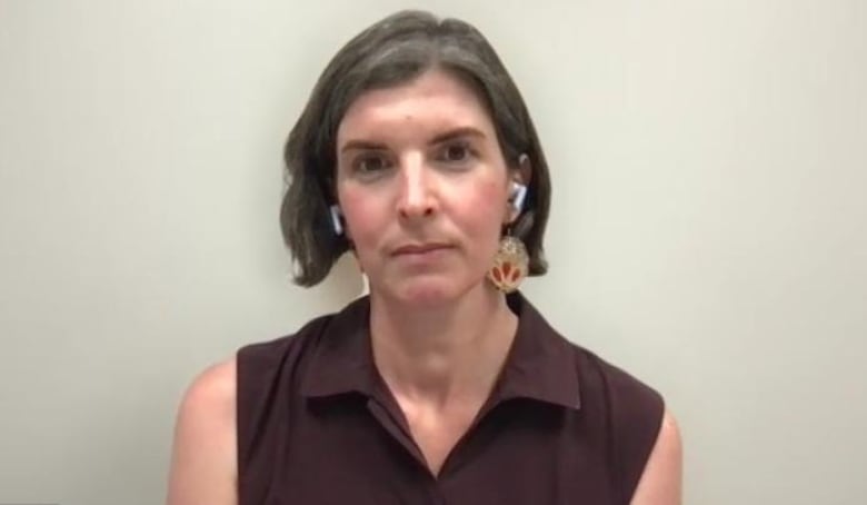 A woman with chin-length brown hair is wearing large gold earrings and a brown sleeveless blouse. She's looking directly at the camera.