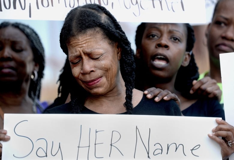 A person cries while holding up a sign that reads, 'Say her name,' as other chant behind them.