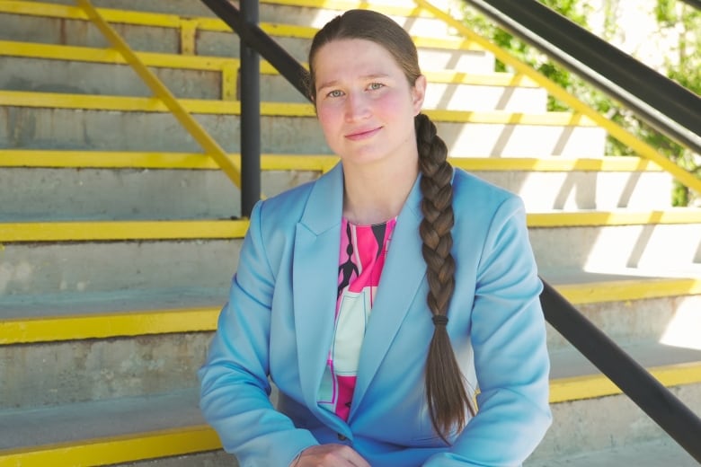 A woman wearing a blue suit-jacket and a pink top underneath it sits on a staircase.