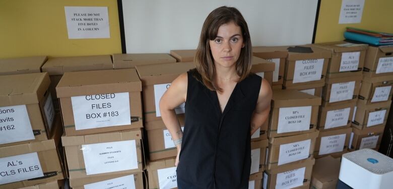 A woman with a serious expression stands in a room in front of stacks of dozens of cardboard file-folder storage boxes.