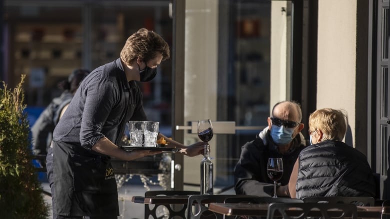 A waiter on an outdoor restaurant patio serves red wine to an older couple dining out on a sunny afternoon in Vancouver. See waiter and couple wearing PPE masks.  Taken on January 22, 2021