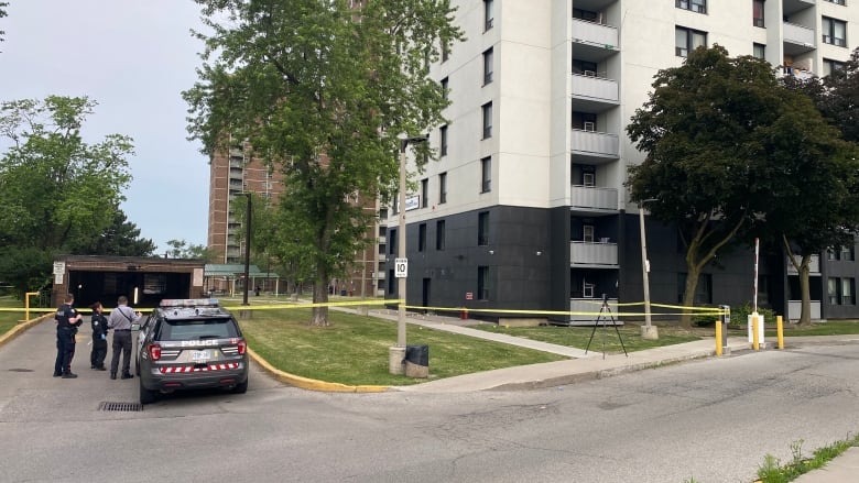 Police huddle around a police car parked outside an apartment building cordoned off with yellow crime scene tape.