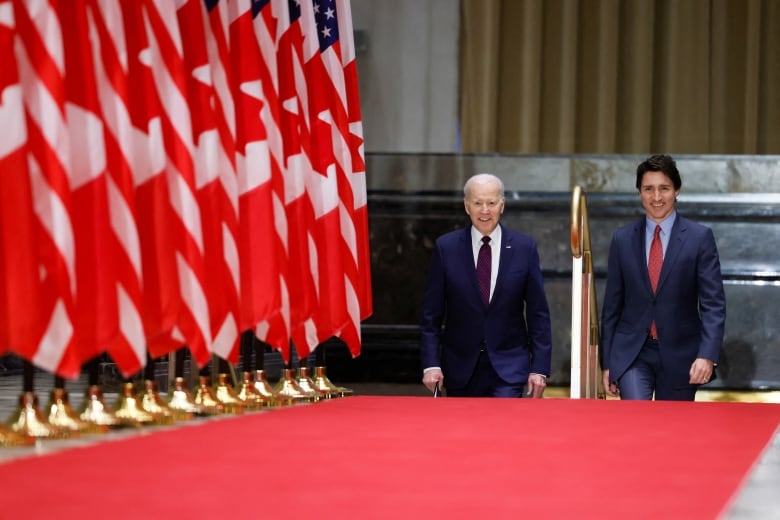 Two politicians halfway up a small staircase leading to a red carpet and hallway lined with flags.