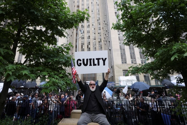A man in a black shirt holds up an American flag and a white sign that says "GUILTY" in black letters in New York City.