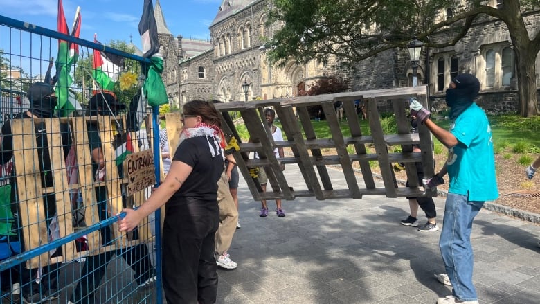 Students on a university campus open a gap in a temporary fence to carry an apple crate out of an encampment on a sunny day