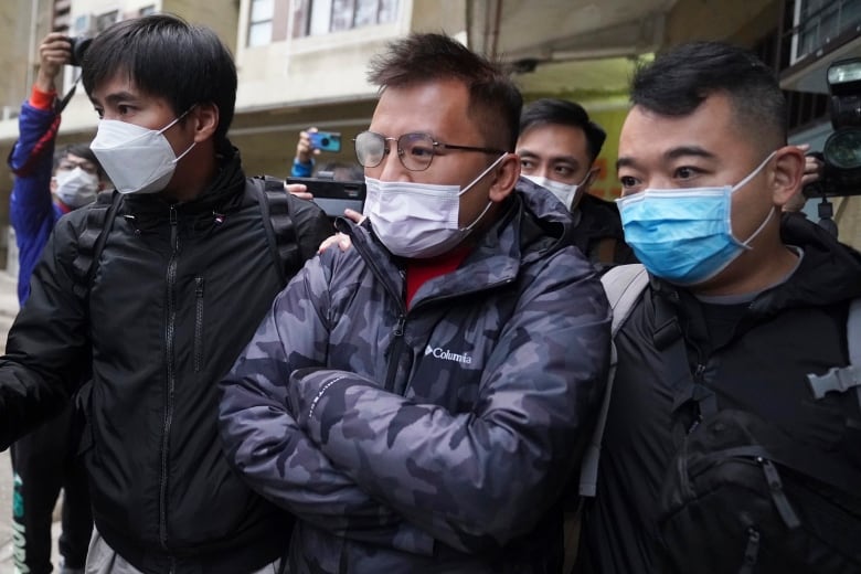 A man wearing glasses and a surgical mask is flanked by two police officers.