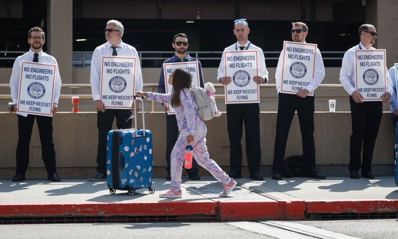 A young female passenger in pink pyjamas makes her way past WestJet airplane mechanics as they stand in a the picket line at Calgary International Airport.