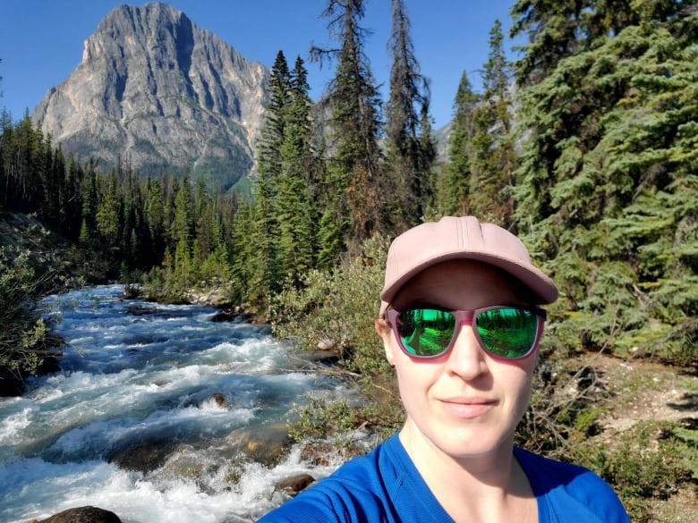 Woman and in sunglasses and hat standing in front of a river and mountain with blue skies in the backdrop.