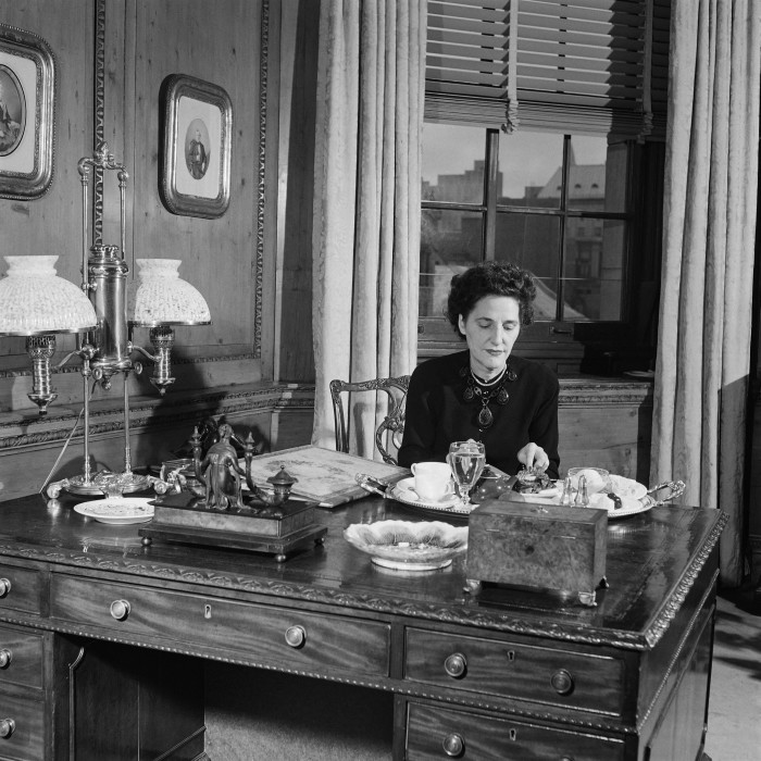 A black-and-white photograph of a woman in a 1940s-style black dress, sits eating at a highly polished wooden desk in an ornately furnished wood-panelled office with heavy curtains behind her